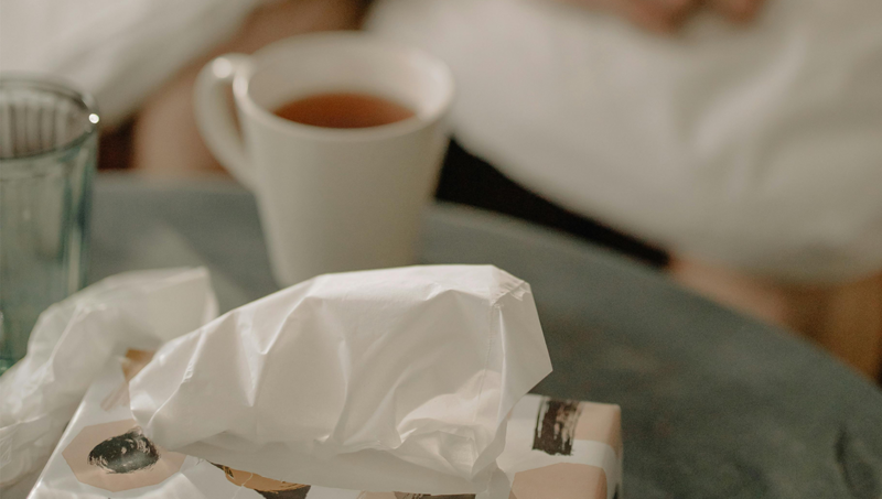 Bild mit Darstellung einer Tschentuchbos und einer Tasse mit Tee auf einem Tisch, sowie unscharf im Hintergrund ein Ausschnitt einer Person. Das Bild soll eine Erkältungssituation darstellen.