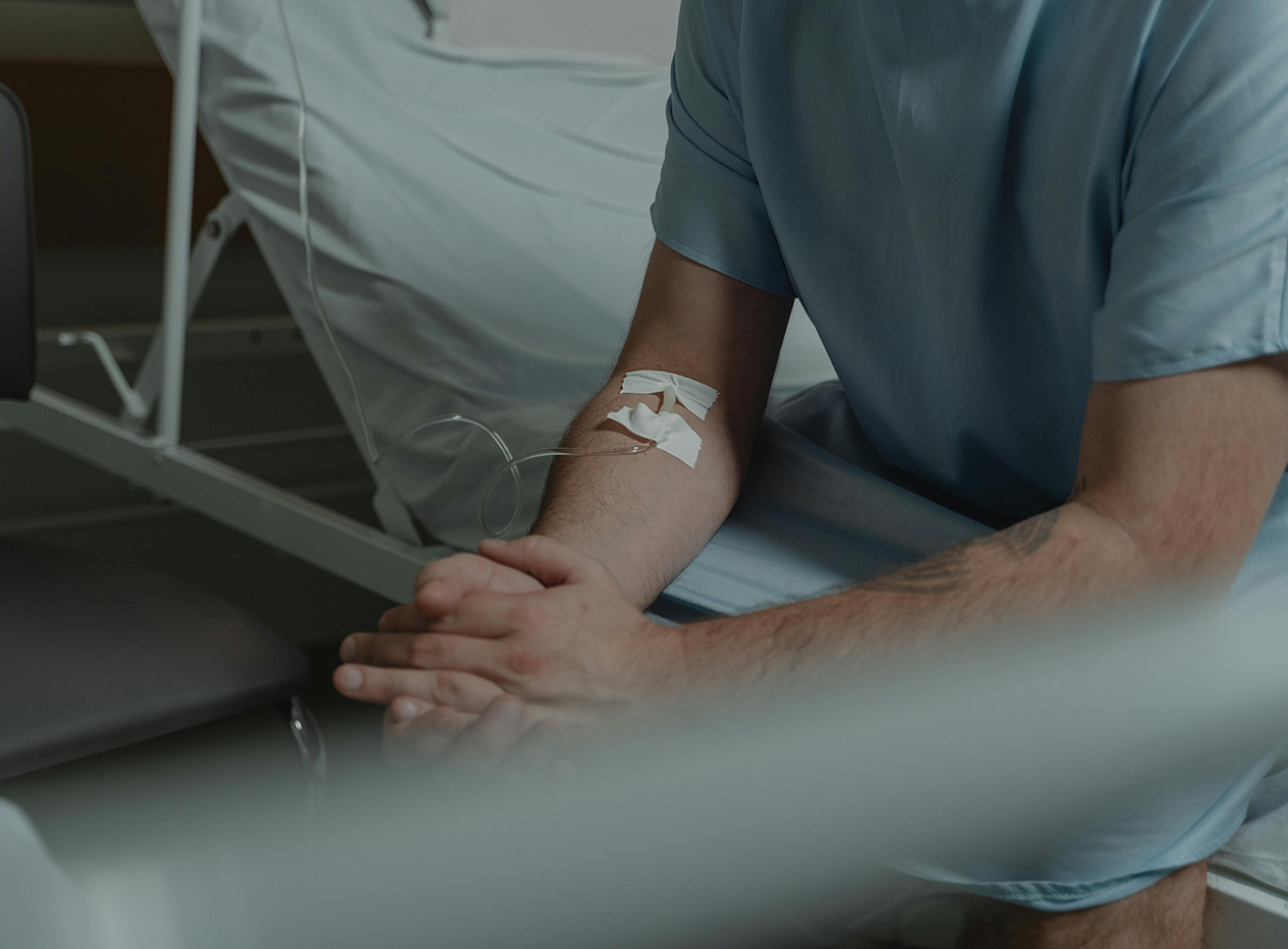 Eine Person sitzt auf einem Patientenbett und hat eine Kanüle in der Armbeuge