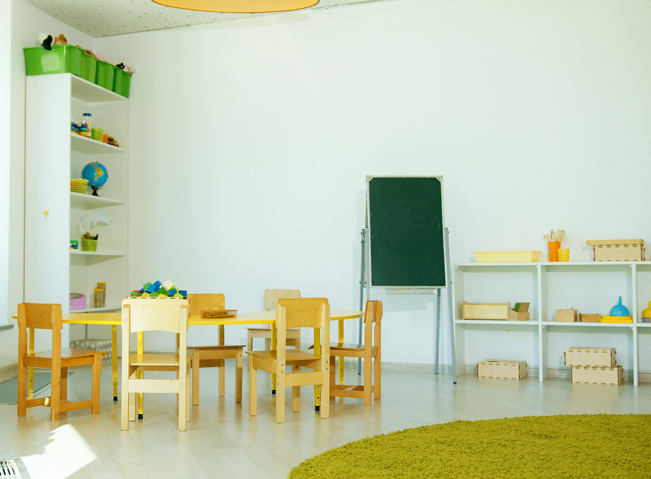 Ein Raum eines Kindergartens in dem ein Tisch mit Stühlen, eine Tafel sowie Regale mit Spielzeug sind.